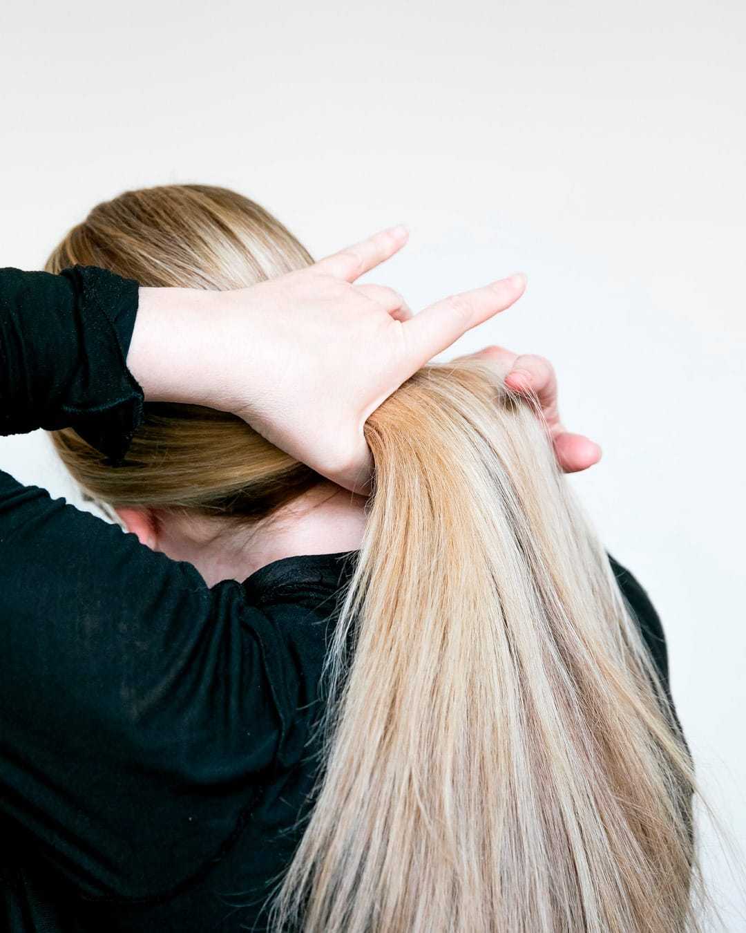 Person styling long blonde hair into a ponytail, wearing a black long-sleeve shirt.