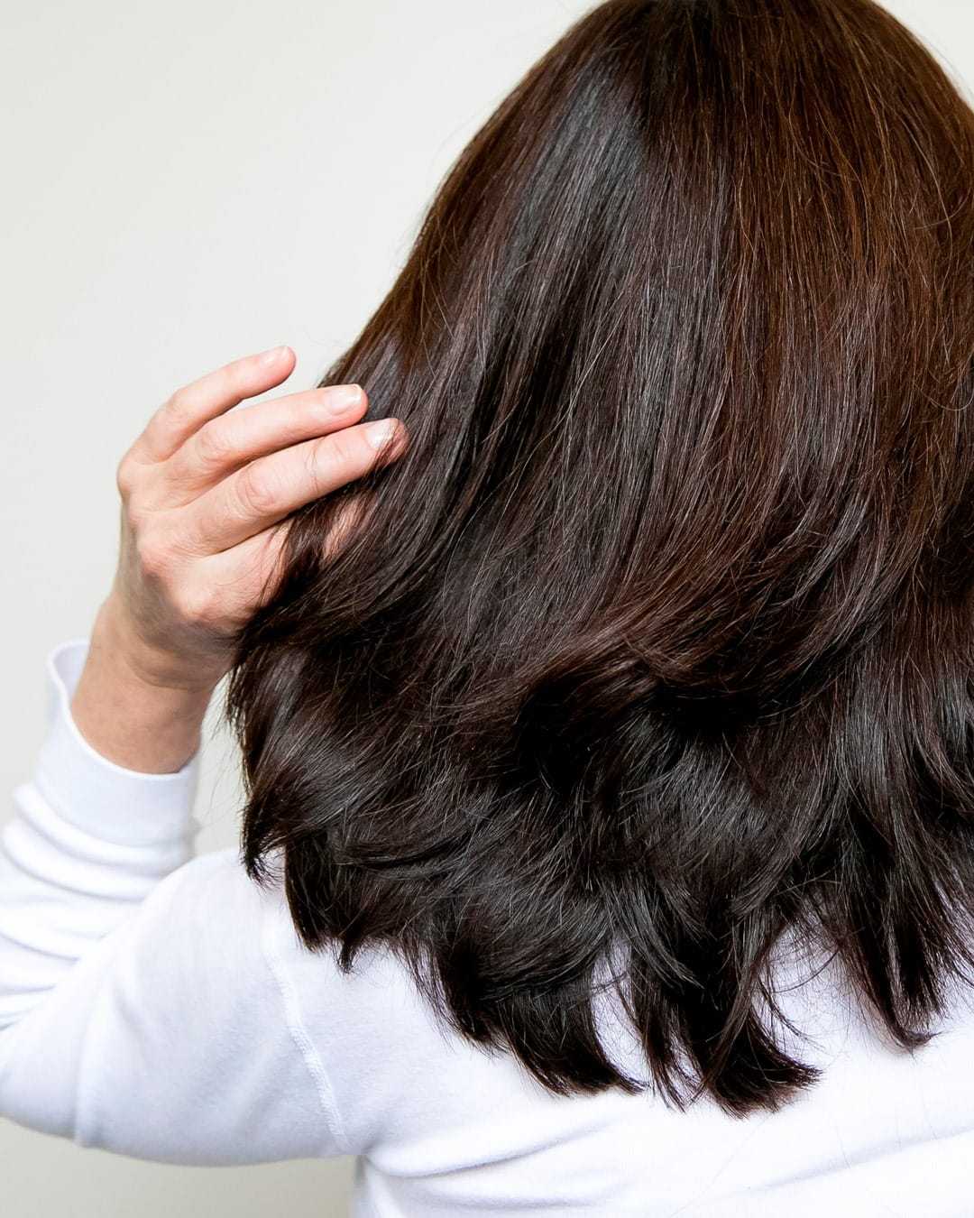 Person with dark, shoulder-length hair touching the back of their head, wearing a white top.