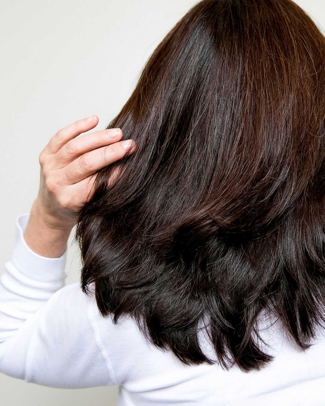 Person with dark, shoulder-length hair touching the back of their head, wearing a white top.