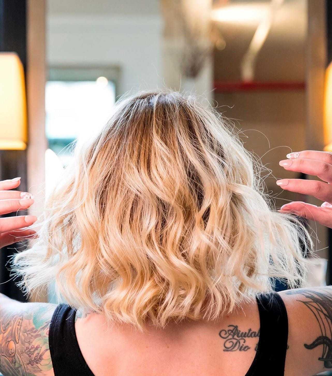 Short, wavy blond hair styled with volume, viewed from the back, with tattooed arms visible.